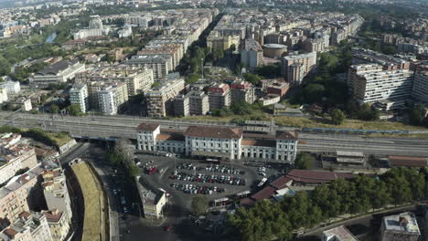 Vista-Aérea-De-Un-Dron-Que-Muestra-La-Estación-De-Tren-Mientras-La-Línea-De-Cercanías-Sale-De-La-Plataforma-En-El-Barrio-De-Trastevere,-Roma,-Italia