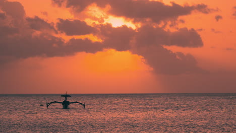 Outrigger-Old-Boat-in-Silhouette-Anchored-Floating-In-Ocean-Against-Orange-Sky-Sunset-During-GOlden-Hour-and-Purple-Clouds