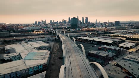 A-retreating-drone-shot-of-the-Ribbon-Of-Light-6th-Street-Bridge-during-sunset-in-Los-Angeles-California-with-cars-driving-in-the-foreground-of-the-city's-skyline,-moving-backwards