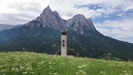 Wunderschöne-Kirche-In-Einem-Tal-Auf-Einer-Grünen-Wiese-Mit-Hohen-Bergen-Im-Hintergrund,-Dolomiten,-Italien,-Europa,-Drohne