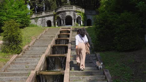 Lady-with-dog-walking-up-stairs-with-flowing-water-in-beautiful-Italian-gardens,-back-view