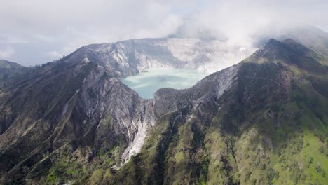 Vista-Aérea-Que-Rodea-El-Borde-De-Un-Volcán-Humeante-Ijen-Con-Un-Lago-Turquesa-Y-Una-Montaña-Nublada-Y-Brumosa-Al-Fondo---Java-Oriental,-Indonesia