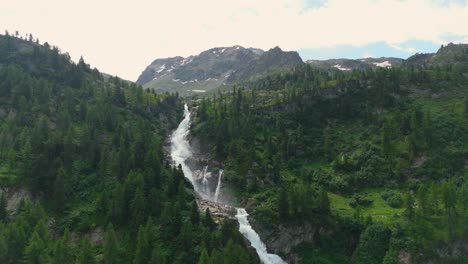 Majestic-waterfall-cascading-down-lush-green-slopes-in-the-Italian-Alps,-aerial-view