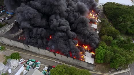 Plumes-of-black-smoke-and-raging-flames-consuming-industrial-warehouse,-aerial