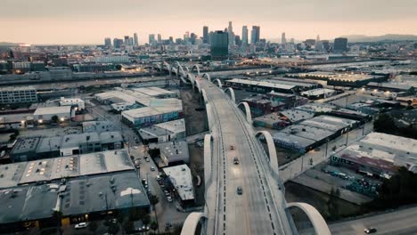 A-drone-shot-of-the-Ribbon-Of-Light-6th-Street-Bridge-during-sunset-in-Los-Angeles-California-with-cars-driving-in-the-foreground-of-the-city's-skyline