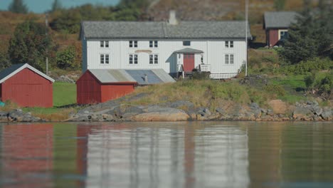 Una-Encantadora-Casa-Blanca-Con-Cobertizos-Rojos-Junto-Al-Agua,-Enmarcada-Por-Un-Exuberante-Paisaje-Verde.