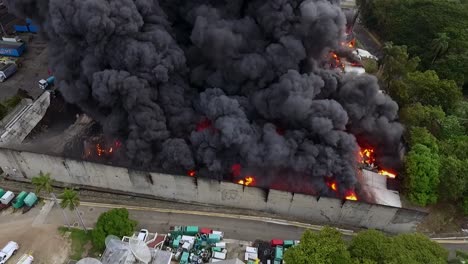 Daytime-warehouse-fire-seen-from-a-drone,-fierce-flames-and-smoke-columns-ascend-into-the-sky-as-emergency-teams-work-to-contain-the-disaster