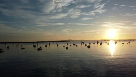 Boats-silhouetted-on-golden-shimmering-harbour-ocean-surface-aerial-view-rising-forwards