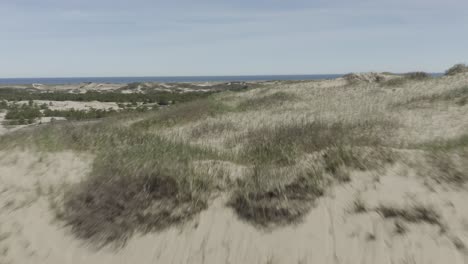 Drone-shot-flying-forwards-over-some-dunes,-revealing-more-dunes-and-the-ocean-behind-it