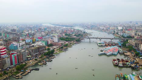 Aerial-view-of-Dhaka-city-featuring-the-bustling-Buriganga-River,-numerous-boats,-and-colorful-buildings-under-a-foggy-sky