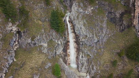 Aerial-view-of-a-vertical-waterfall-plunging-down-rocky-cliffs-in-the-Dolomites,-surrounded-by-alpine-terrain