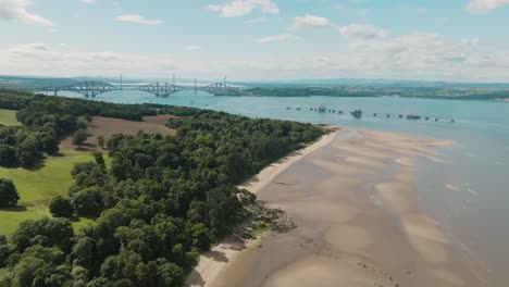 Aerial-view-of-scenic-Scottish-coastline-and-Firth-of-Forth,-yellow-sandy-beach-at-low-tide-next-to-woodland-shoreline-lifting-up-to-reveal-the-famous-South-Queensferry-Bridges,-Scotland,-UK