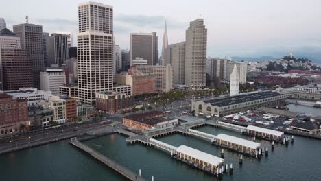 Zoom-out-aerial-camera-shot-from-a-drone-shows-the-Ferry-Building-along-the-San-Francisco,-showcasing-historic-building’s-architecture-against-the-busy-Embarcadero-and-Bay-Bridge