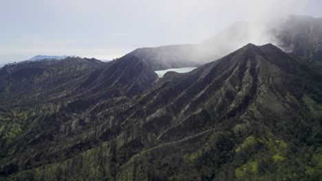 Aerial-Closeup-of-the-rim-of-a-steaming-volcano-Ijen-with-a-Turquoise-Lake,-and-foggy-cloudy-Mountain-in-the-background---East-Java,-Indonesia