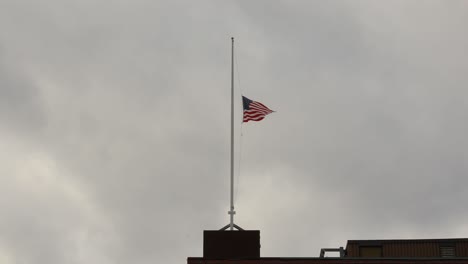 Una-Fuerte-Brisa-Hace-Ondear-La-Bandera-De-Los-Estados-Unidos-A-Media-Asta-Contra-Un-Cielo-Gris-Tormentoso-En-4k