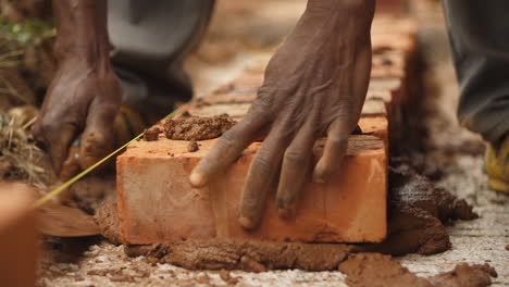 Primer-Plano-De-Un-Constructor-Africano-Colocando-Ladrillos-Y-Recogiendo-Una-Mezcla-De-Cemento-Marrón-Con-Una-Paleta