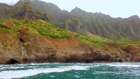 4K-Hawaii-Kauai-Boating-on-ocean-left-to-right-pan-past-waterfall-along-rocky-shoreline-with-mountains-in-cloudy-distance