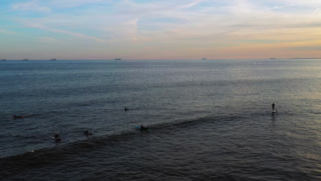 low-altitude-drone-camera-hovers,-then-pan-right-watching-the-surfers-as-they-wait-for-the-perfect-wave-and-a-golden-sunset-in-the-background