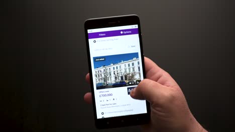 Nahaufnahme-Einer-Männlichen-Hand,-Die-Mit-Einem-Smartphone-Durch-Immobilienanzeigen-Auf-Einer-Englischen-Website-Scrollt