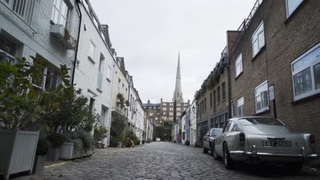 Scenic-wide-low-angle-view-of-cobblestone-narrow-street-and-old-fancy-car-parked-by-urban-brick-building,-London,-England,-handheld-pan-right-slow-motion