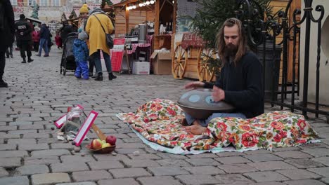 Long-haired-street-musician-playing-Hang-drum-on-market-square-to-earn-money