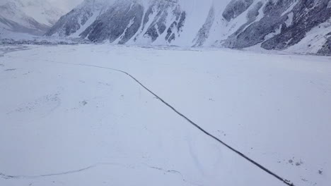 Aerial-Shot-of-Car-driving-down-a-single-snow-covered-road-with-snowy-mountains-in-the-background