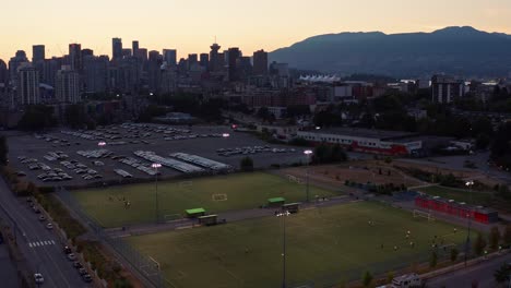 Vista-Aérea-De-Un-Dron-Al-Atardecer-En-El-Norte-De-Vancouver-Sobre-Campos-De-Fútbol:-Puesta-De-Sol-Detrás-Del-Paisaje-Urbano-De-Columbia-Británica,-Canadá