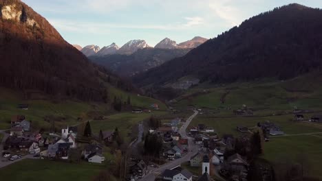 Golden-alpenglow-high-above-the-shadowed-village-of-Stein-SG,-Toggenburg,-Switzerland,-aerial-establish