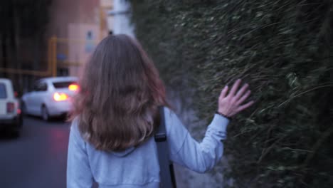 Beautiful-curly-haired-girl-walks-along-the-road-and-touches-the-hedge-hanging-from-the-fence,-hedgerow,-following-after-a-person-from-behind