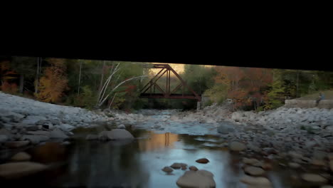 Toma-Panorámica-Desde-La-Superficie-Quieta-Del-Río-Debajo-Del-Puente-Viejo,-Que-Revela-Montañas-Y-Follaje.