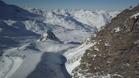 Ski-descent-of-Val-Thorens-ski-resort-next-to-jagged-rocky-mountain-slope,-in-the-French-Alps---Aerial-ascending-Fly-over-shot