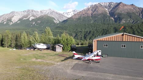 Rotating-aerial-shot-of-a-Cessna-206,-aircraft-hanger-and-the-Talkeetna-Mountain-range-at-a-private-grass-airfield