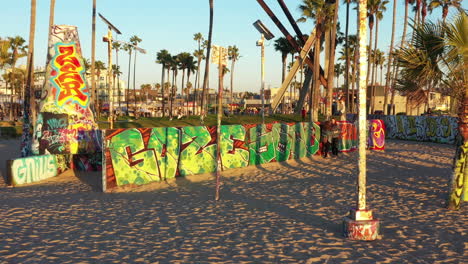 Drone-shot-of-venice-beach-boardwalk-during-sunset-showing-palm-trees,-graffiti-walls,-skateboarding-and-people-taking-photos