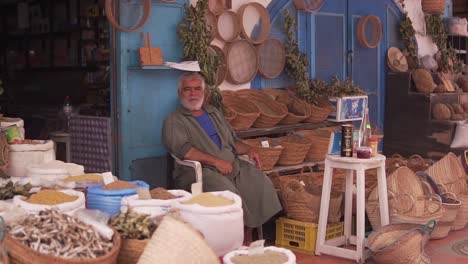 Un-Anciano-En-La-Marchandise-De-Djerba-Houmet-Souk-Vendiendo-Accesorios-Y-Decoraciones-Frente-A-Su-Tienda