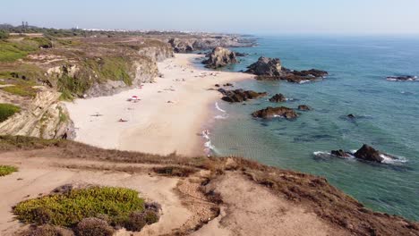 Praia-da-Samoqueira-Beach-near-Porto-Covo,-Alentejo,-West-Portugal---Aerial-Drone-View-of-motorhomes-,-golden-sandy-beach-and-rocky-coastline