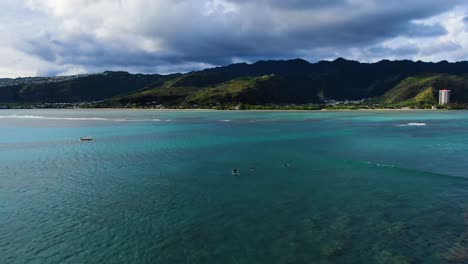 Four-male-surfers-sitting-on-their-surfboards-while-waiting-for-waves-far-away-from-shore-with-green-mountain-range-in-the-background