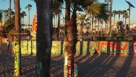 Drone-shot-of-venice-beach-boardwalk-during-sunset-showing-palm-trees,-graffiti-walls,-skateboarding-and-people-taking-photos