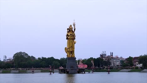 Close-static-shot-of-golden-Lord-Shiva-statue-centered-in-Sursagar-Lake-on-cloudy-day-in-Vadodara,-Gujarat,-India-with-ample-copy-space