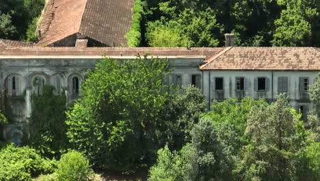 Overgrown-abandoned-school-building-in-Italy,-surrounded-by-lush-green-trees,-daytime,-aerial-view