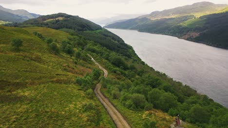 Aerial-view-of-Scotland,-bagpiper-playing-on-a-mountain-overlooking-loch-in-the-Scottish-highlands,-United-Kingdom