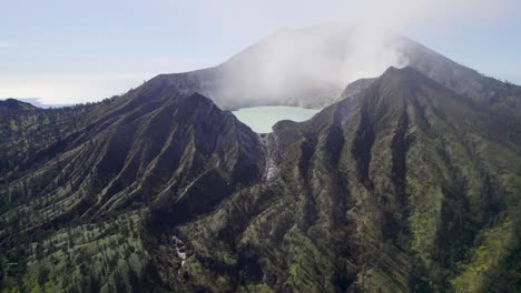 Aerial-Closeup-of-the-rim-of-a-steaming-volcano-Ijen-with-a-Turquoise-Lake,-and-foggy-cloudy-Mountain-in-the-background---East-Java,-Indonesia