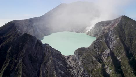 Aerial-Closeup-of-the-rim-of-a-steaming-volcano-Ijen-with-a-Turquoise-Lake,-and-foggy-cloudy-Mountain-in-the-background---East-Java,-Indonesia