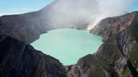 Aerial-Closeup-of-the-rim-of-a-steaming-volcano-Ijen-with-a-Turquoise-Lake,-and-foggy-cloudy-Mountain-in-the-background---East-Java,-Indonesia