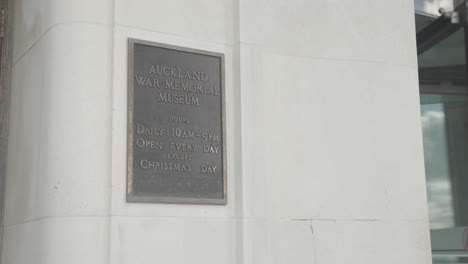 Still-view-of-Auckland-War-Memorial-Museum-plaque-on-the-wall-with-open-hours,-near-a-spinning-turnstile