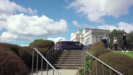 People-walk-down-the-steps-that-lead-to-the-Auckland-War-Memorial-Museum-near-bushes-and-grass-on-a-sunny-day-with-clouds-in-the-sky