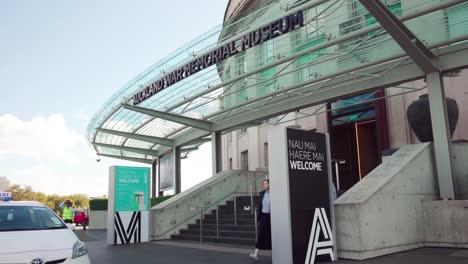 A-couple-exits-the-Auckland-War-Memorial-Museum-and-walks-by-two-welcome-signs-on-a-sunny-day-in-New-Zealand