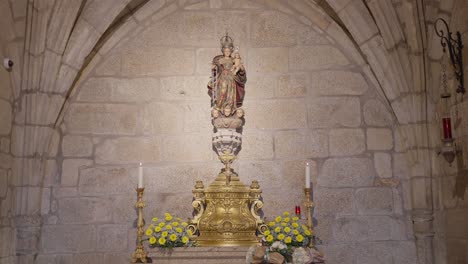 Statue-of-the-Virgin-Mary-with-Child-on-a-golden-altar-in-a-stone-church,-surrounded-by-flowers-and-candles,-symbolizing-devotion-and-religious-heritage