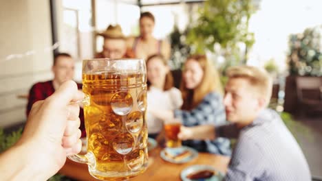 Focus-on-hand-holding-mug-with-lager-beer.-Group-of-young-people,-friends-meeting-together,-drinking-beer,-having-fun.-Concept-of-oktoberfest,-traditional-taste,-friendship,-leisure-time,-enjoyment