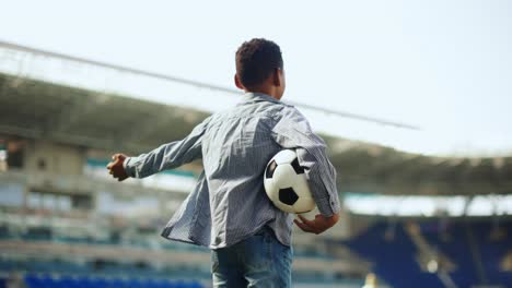 Little-boy,-child-standing-at-empty-sport-stadium-with-football-ball-and-spreading-hand.-Dreaming-of-becoming-soccer-sportsman.-Concept-of-childhood,-dreams,-imagination-and-future,-sport