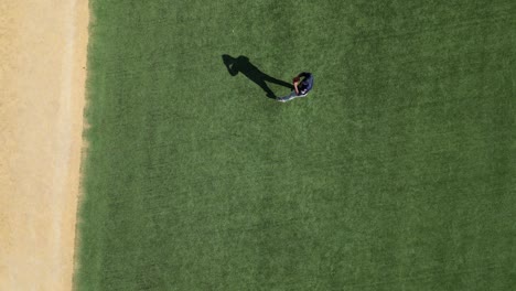 A-drone-shot-of-a-baseball-player-throwing-and-receiving-the-ball-on-green-lawn-pitch